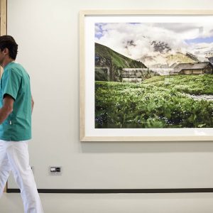 A woman wearing a yellow jacket, hat, and backpack stands on a rocky ledge, looking out over a wide river and forested valley below, with mountains visible in the distance under a cloudy sky.