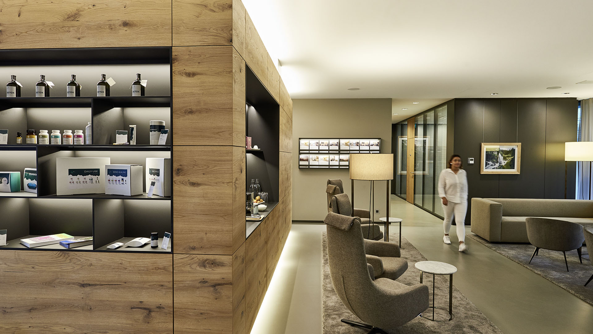 A modern, well-lit lounge features wooden shelves displaying products, comfortable chairs, and a woman in white walking through the space. Warm lighting and minimalist decor create a welcoming atmosphere.